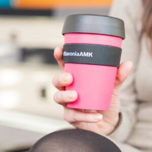 Beigeen puseroon pukeutunut henkilö pitelee vaaleanpunaista uudelleenkäytettävää kahvikuppia, jossa on musta kansi ja nauha. Nauhaan on painettu teksti SavoniaAMK. Tausta on sumea.