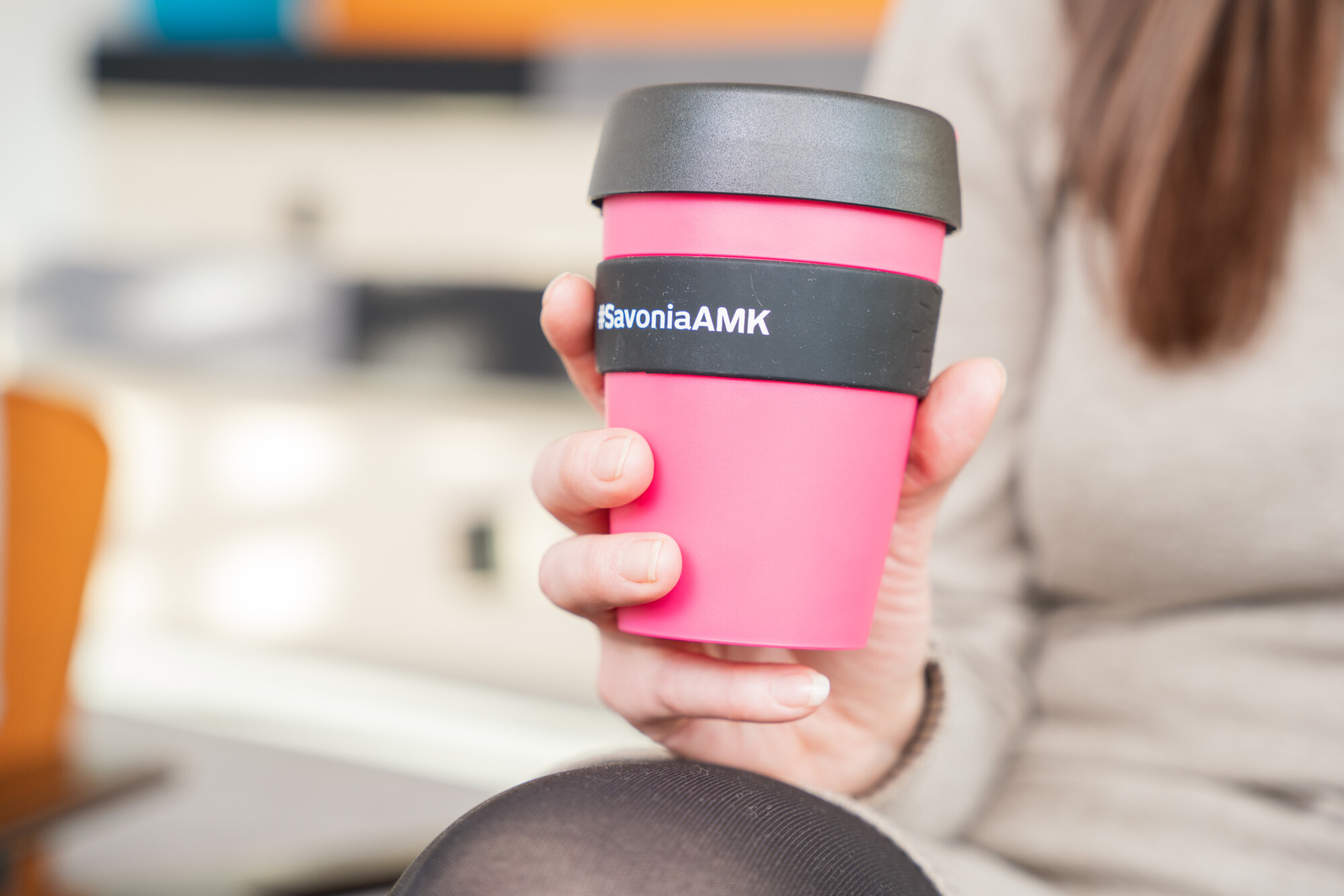 Beigeen puseroon pukeutunut henkilö pitelee vaaleanpunaista uudelleenkäytettävää kahvikuppia, jossa on musta kansi ja nauha. Nauhaan on painettu teksti SavoniaAMK. Tausta on sumea.