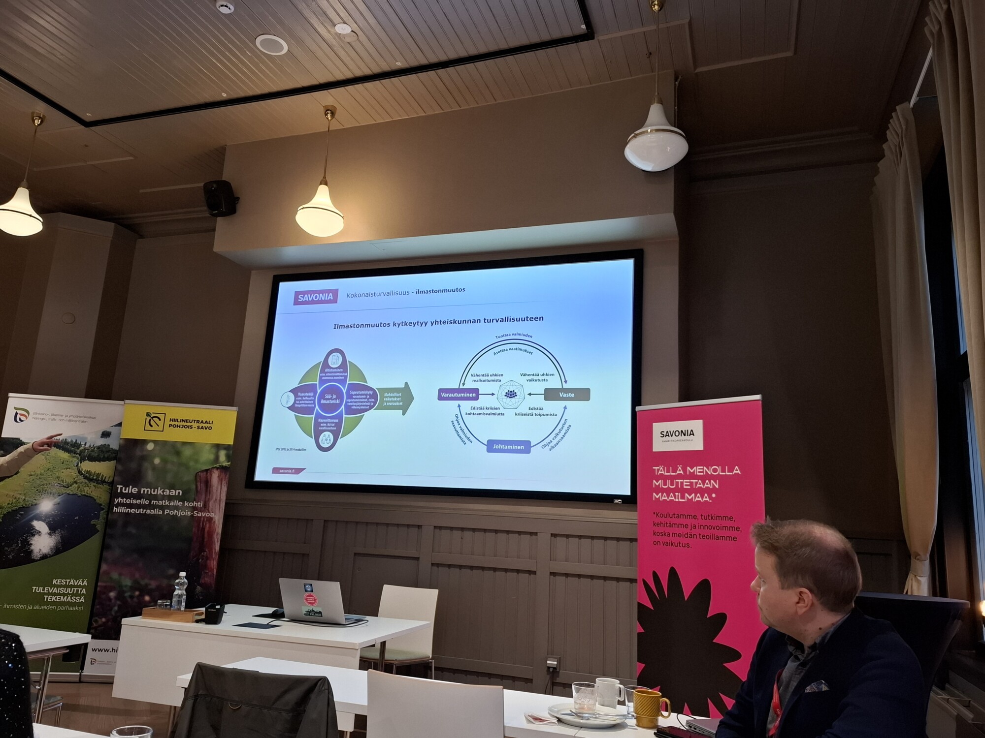 Konferenssihuone, jossa on valkokankaalle heijastettu esitys. Ihmiset istuvat pöydän ääressä kannettavien tietokoneiden ja kahvin kanssa. Bannereita ja tiedotusjulisteita on esillä ympäri huonetta.