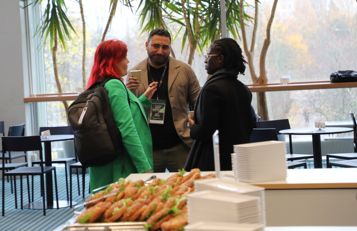 Kolme ihmistä seisoo ja puhuu valoisassa huoneessa, jossa on suuret ikkunat ja kasveja. Yhdellä naisella on vihreä takki ja reppu, kun taas kahdella miehellä on bleiserit. Etualalla näkyy tarjottimella olevia voileipiä ja pinottuja lautasia.