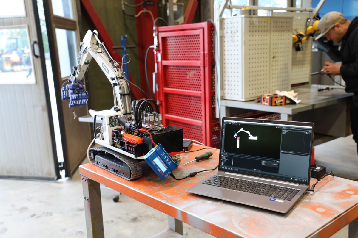 Pieni robottikaivinkone istuu työpöydällä kannettavan tietokoneen vieressä, jossa on sen käyttöliittymä. Työkaluja ja laitteita on ripoteltu ympäriinsä, ja taustalla työskentelee henkilö työpajan ympäristössä.
