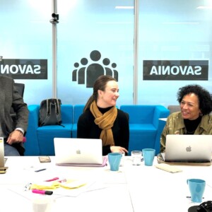 Kolme ihmistä istuu pöydän ääressä kannettavien tietokoneiden kanssa, keskustelevat ja hymyilevät. Heidän takanaan on sinisiä sohvia ja suuria ikkunoita, joihin on painettu sana "SAVONIA". Ympäristö vaikuttaa modernilta toimisto- tai kokoushuoneelta.