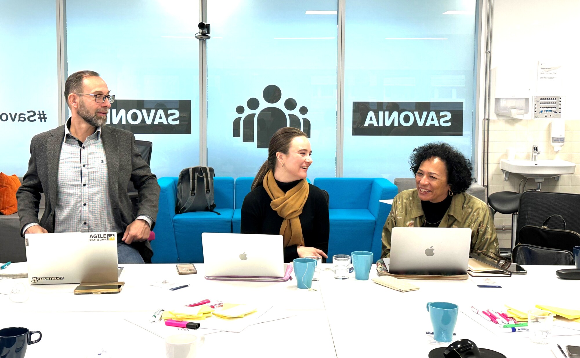 Kolme ihmistä istuu pöydän ääressä kannettavien tietokoneiden kanssa, keskustelevat ja hymyilevät. Heidän takanaan on sinisiä sohvia ja suuria ikkunoita, joihin on painettu sana 