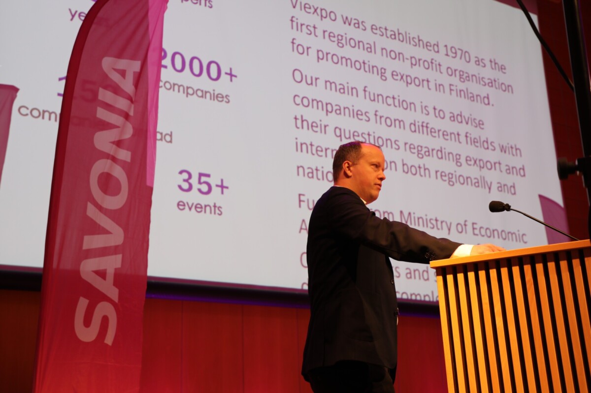 Pukumies puhuu lavalla korokkeella vaaleanpunaisen SAVONIA-banderollin vieressä. Hänen takanaan olevalla suurella näytöllä näkyy tekstiä Suomen vientiä edistävästä järjestöstä.