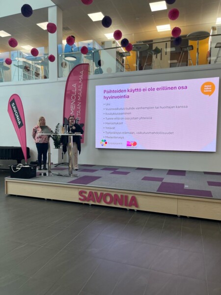 Kaksi naista seisoo lavalla mikrofonien kanssa Savonia-banderollin vieressä. Heidän takanaan olevalla suurella näytöllä näkyy suomenkielinen dia päihteiden käytön vaikutuksista hyvinvointiin. Tapahtumapaikka on koristeltu vaaleanpunaisilla ja valkoisilla roikkuvilla palloilla.