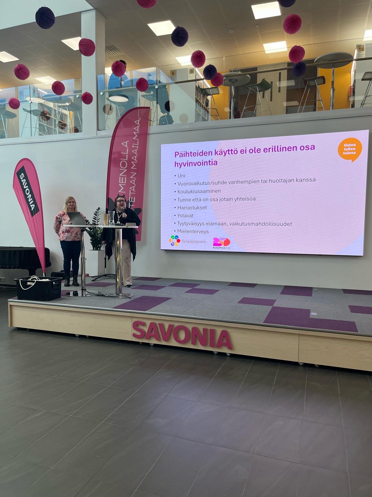 Kaksi naista seisoo lavalla mikrofonien kanssa Savonia-banderollin vieressä. Heidän takanaan olevalla suurella näytöllä näkyy suomenkielinen dia päihteiden käytön vaikutuksista hyvinvointiin. Tapahtumapaikka on koristeltu vaaleanpunaisilla ja valkoisilla roikkuvilla palloilla.