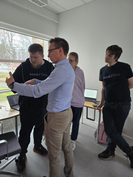 Vaaleansiniseen paitaan pukeutunut mies tekee eleitä puhuessaan kolmelle henkilölle, joista kahdella on koodin / Sisun T-paita, huoneessa, jossa on kannettavat tietokoneet ja suuret ikkunat. He näyttävät keskustelevan tai työskentelevän yhdessä.