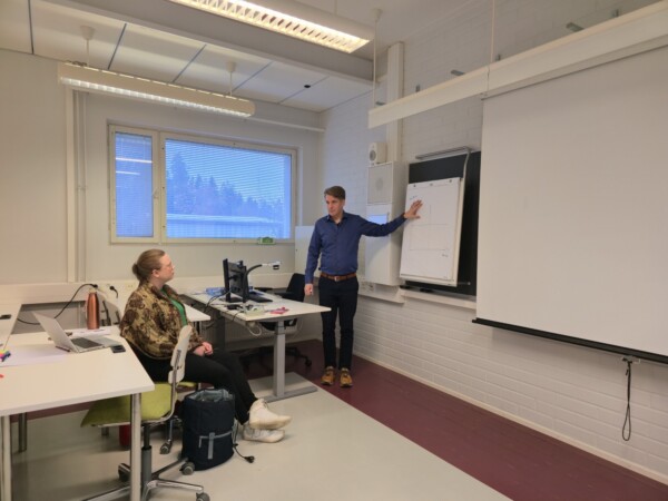 Mies seisoo ja osoittaa fläppitaulua luokkahuoneessa pidettävän esityksen aikana, kun taas nainen istuu pöydän ääressä kannettavan tietokoneen kanssa ja kuuntelee tarkkaavaisesti.