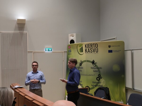 Kaksi miestä seisoo luentosalin edessä puhumassa yleisölle. Heidän takanaan on vihreä Kierto Kasvu -näyttö, jossa on tietoa kestävästä kehityksestä ja kierrätyksestä. Tuolit ja osa yleisöstä ovat näkyvissä.
