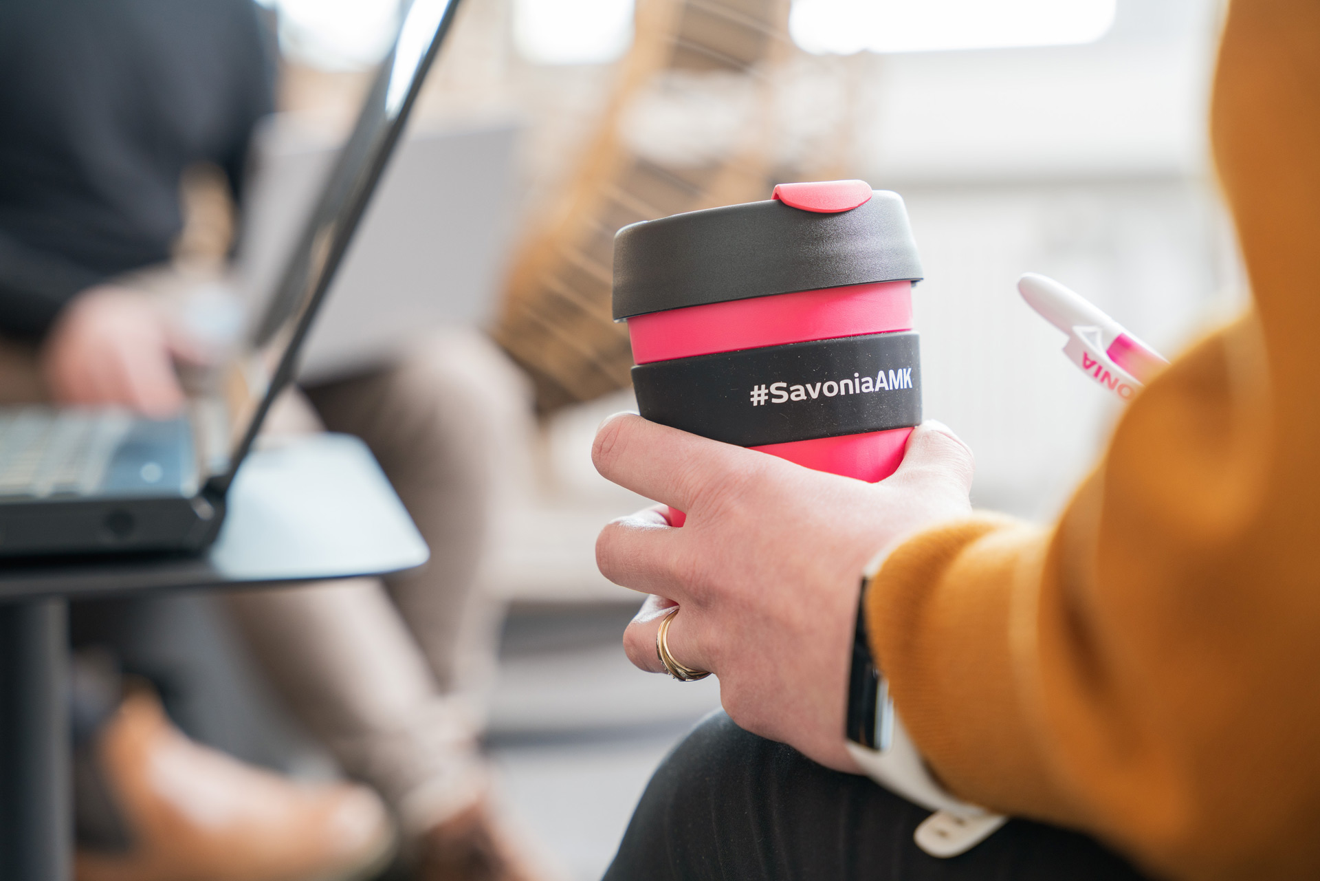 Keltaiseen villapaitaan pukeutunut henkilö pitää kädessään vaaleanpunaista uudelleenkäytettävää mukia, jossa lukee #SavoniaAMK, ja kynää kannettavan tietokoneen vieressä. Sumeassa taustalla toinen henkilö käyttää kannettavaa tietokonetta.