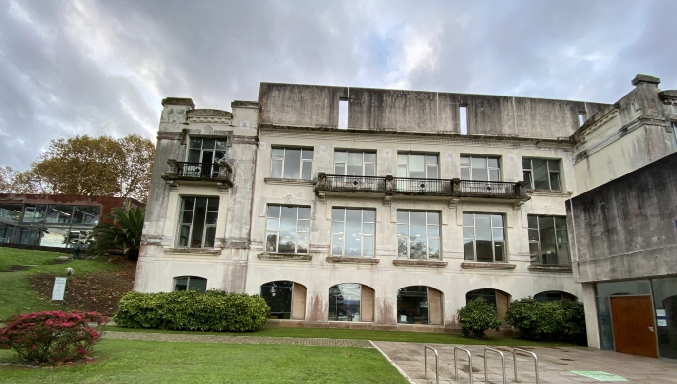 Suuri, valkoinen, kolmikerroksinen rakennus, jossa on parvekkeita ja paljon ikkunoita, ja jossa on iän ja sään merkkejä. Rakennusta ympäröivät vihreät pensaat, ruoho ja polkupyöräteline, ja yläpuolella on pilvinen taivas.