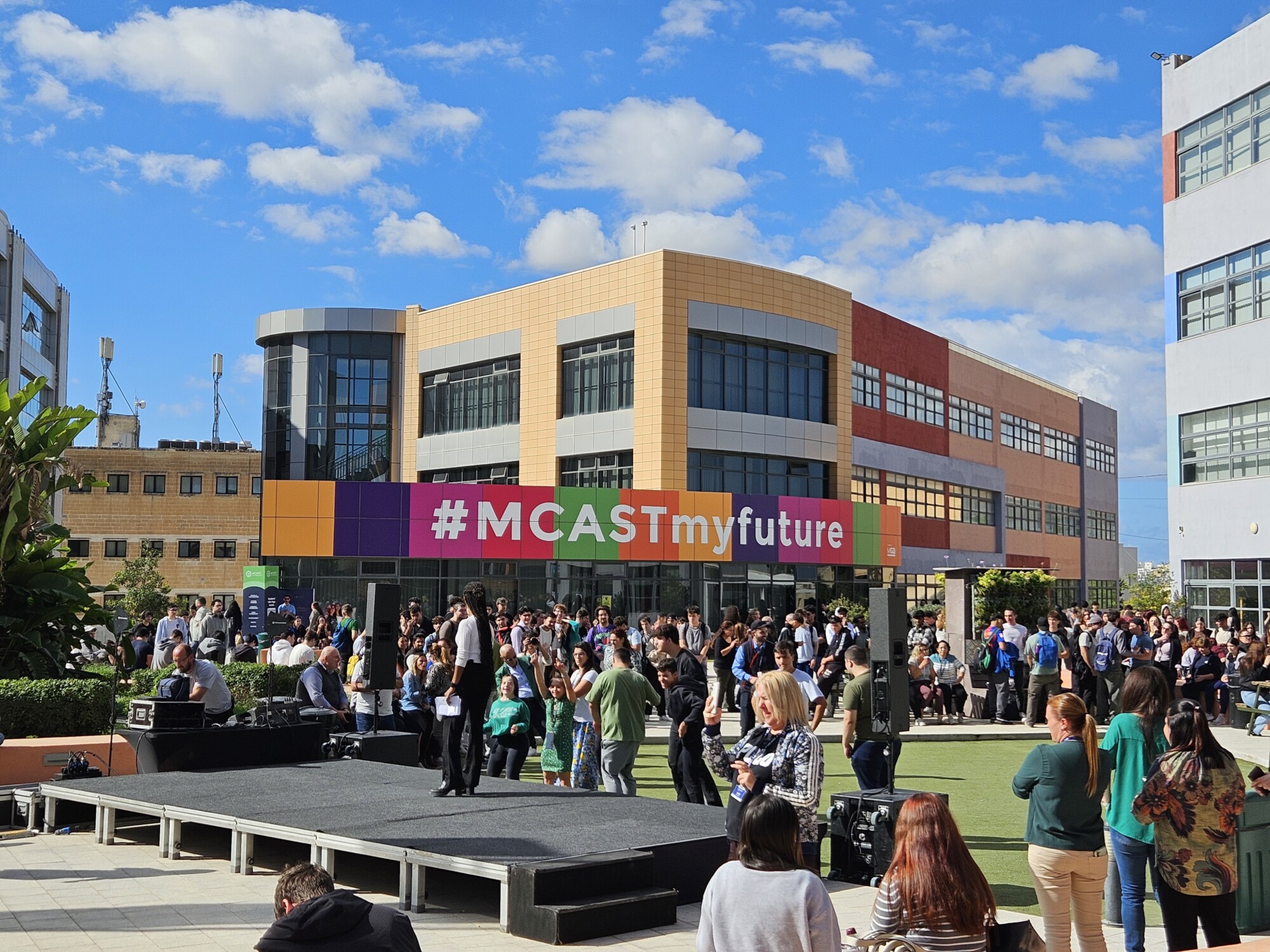 Ihmisjoukko kokoontuu modernin rakennuksen ulkopuolelle, jonka julkisivussa on suuri #MCASTmyfuture-banneri. Ihmiset seisovat lavan ympärillä kirkkaan sinisellä taivaalla, jolla on hajapilviä.
