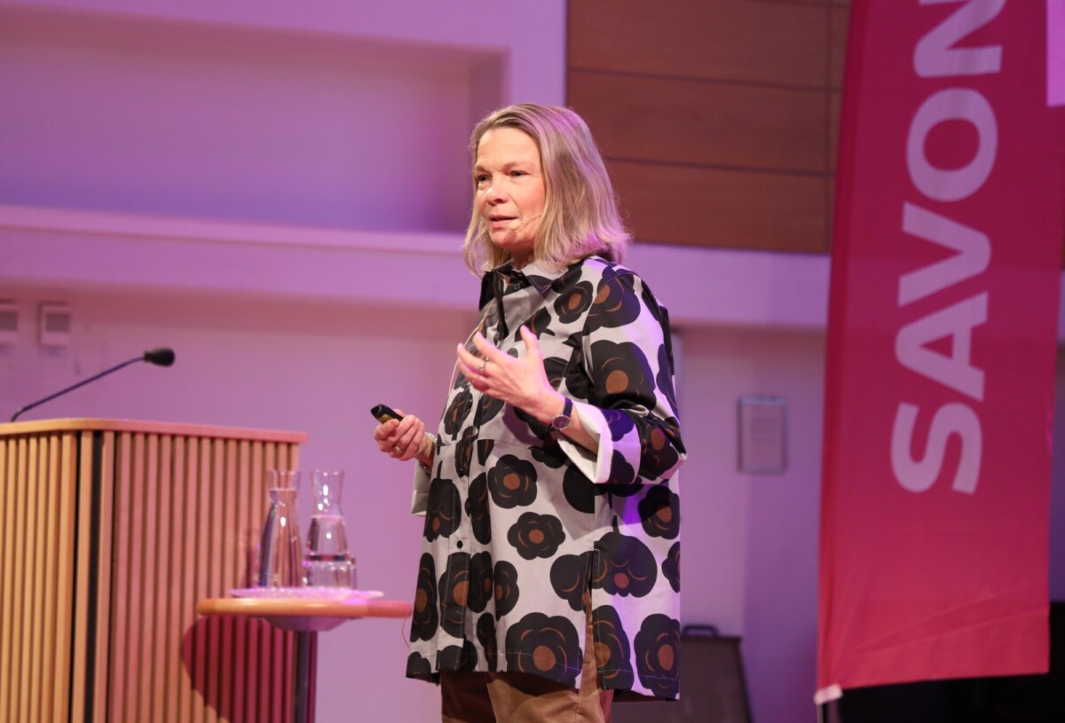Lavalla puhuu nainen, jolla on olkapäiden pituiset vaaleat hiukset, joka elehtii toisella kädellään ja pitää toisessa kädessään pientä laitetta. Hän seisoo lähellä pulpettia, jossa on lasisia kannuja. Taustalla näkyy suuri punainen banderolli, jossa lukee SAVON.