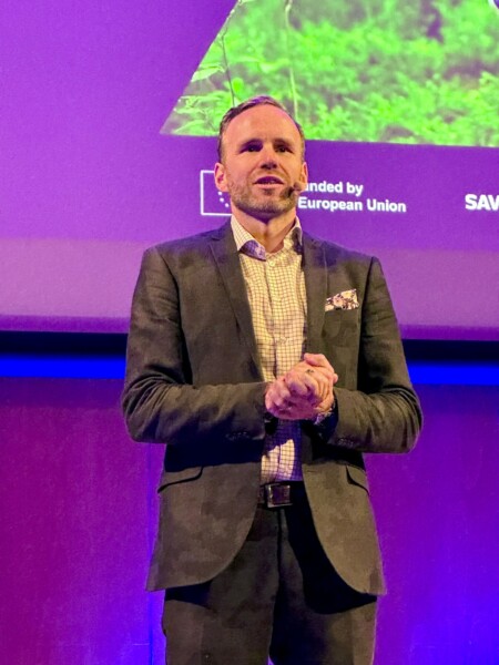 Pukumies seisoo lavalla Euroopan unionin tekstiä esittävän violetin ruudun edessä ja puhuu yleisölle kädet ristissä.