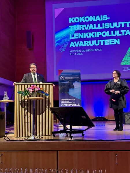 Pukumies puhuu korokkeella, jossa on kukkia lavalla, ja mustaan kaapuun pukeutunut nainen seisoo sivussa. Heidän takanaan on suuri näyttö, jossa on suomenkielistä tekstiä ja julisteen näyttö.