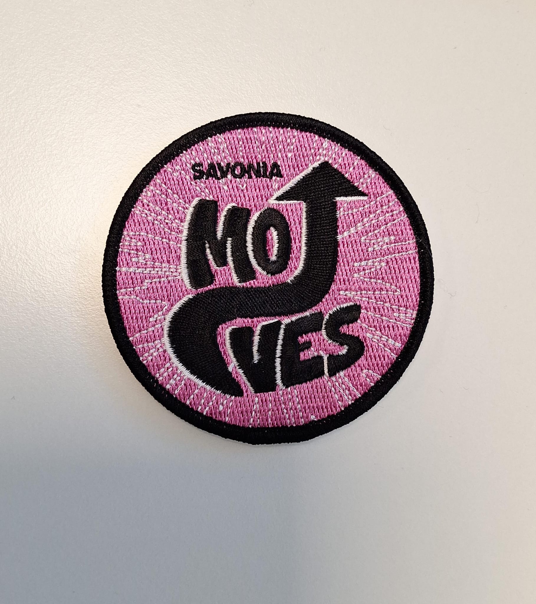 Pyöreässä vaaleanpunaisessa laastarissa, jossa on musta reunus, lukee mustalla sana MOVES, jossa O ja V muodostavat ylöspäin osoittavan kaarevan nuolen. Yläpuolella on mustalla kirjailtu sana SAVONIA.