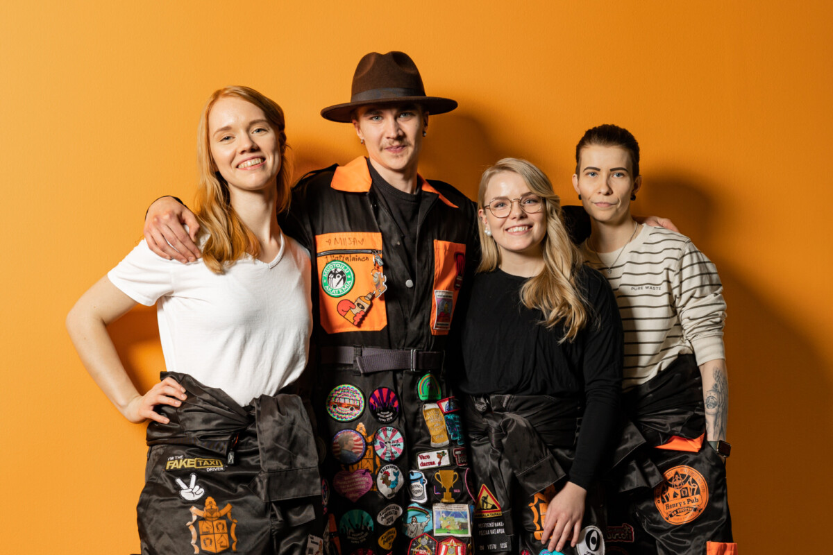 Neljä ihmistä seisoo yhdessä hymyillen oranssin seinän edessä. Heillä on yllään mustat haalarit, joita koristavat värikkäät laastarit, ja yhdellä henkilöllä on ruskea hattu ja oranssi paita.