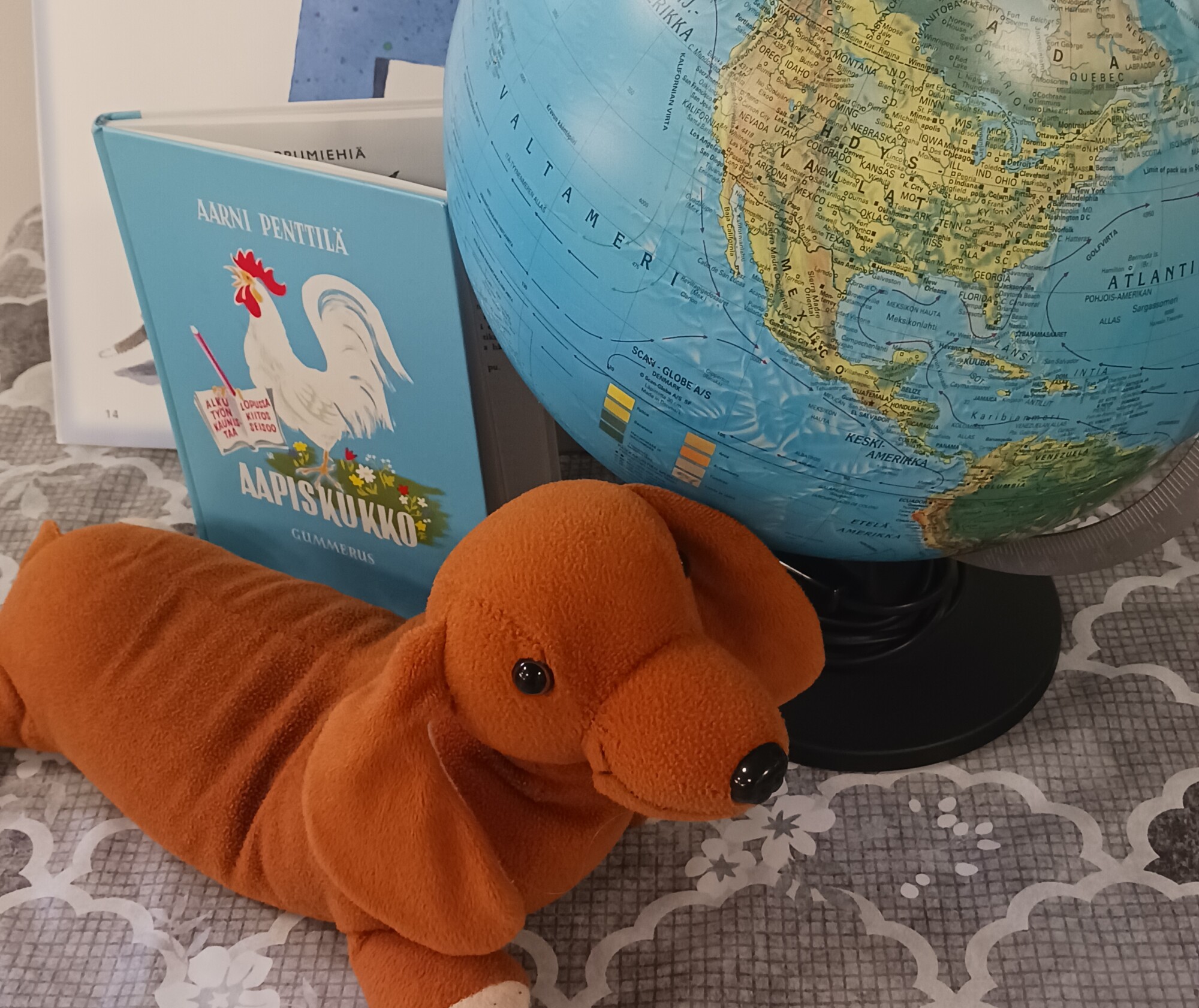 Ruskea muhkea mäyräkoiran lelu makaa kuvioidulla pinnalla Pohjois-Amerikkaa esittävän maapallon vieressä. Niiden takana on sininen suomalainen ABC-kirja, jonka kannessa on kukkokuvitus.