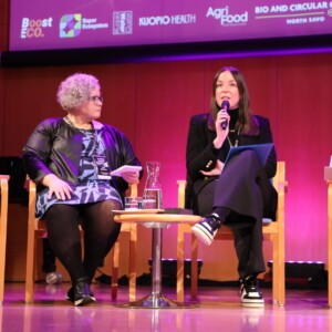 Lavalla istuu neljä ihmistä, jotka käyvät paneelikeskustelua. Kolmella naisella ja yhdellä miehellä on kädessään mikrofonit tai muistiinpanot. He istuvat tuoleilla, joiden edessä on pyöreä pöytä, ja heidän takanaan olevalla ruudulla näkyy tapahtuman sponsoreiden logoja.