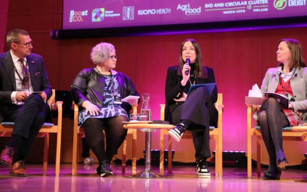 Lavalla istuu neljä ihmistä, jotka käyvät paneelikeskustelua. Kolmella naisella ja yhdellä miehellä on kädessään mikrofonit tai muistiinpanot. He istuvat tuoleilla, joiden edessä on pyöreä pöytä, ja heidän takanaan olevalla ruudulla näkyy tapahtuman sponsoreiden logoja.