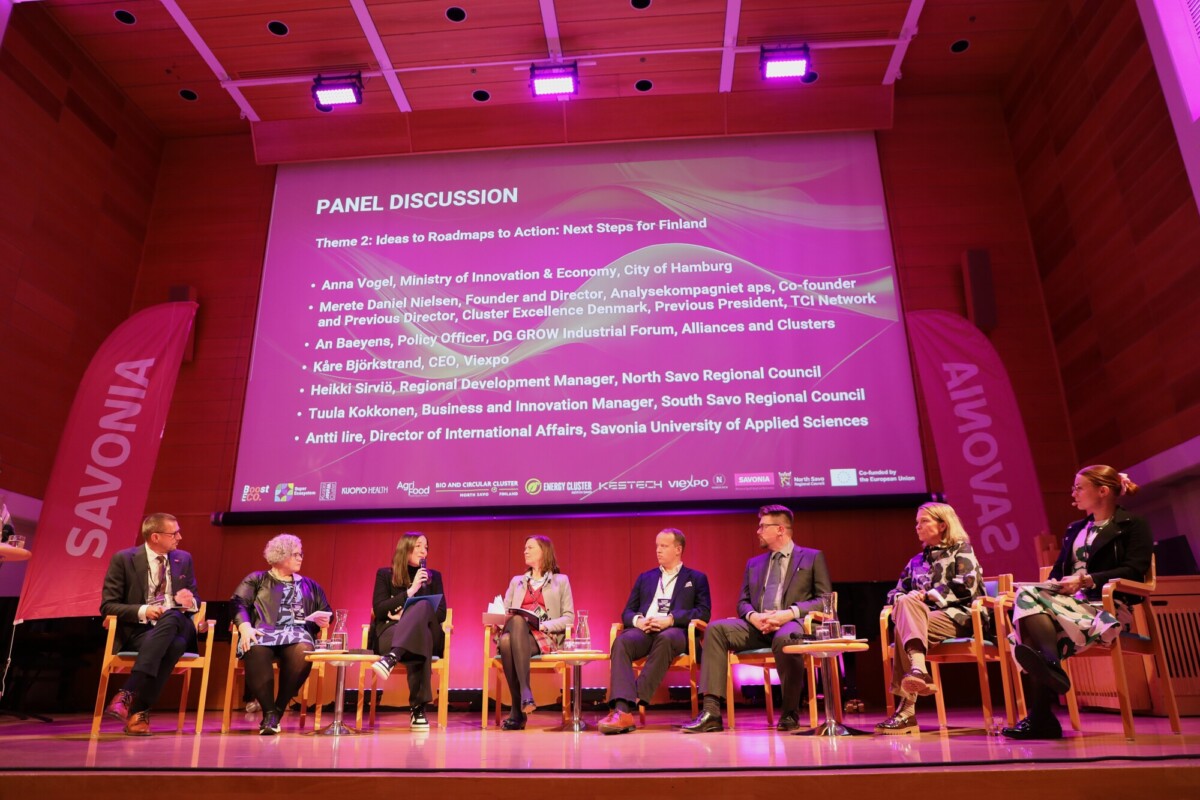 Kahdeksan panelistia istuu lavalla, kun heidän nimensä ja tittelinsä näkyvät valkokankaalla konferenssin paneelikeskustelun aikana. Lava on valaistu violeteilla valoilla, ja taustalla lukee SAVONIA.