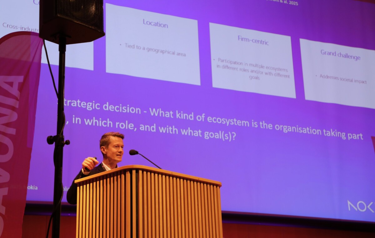 Mies seisoo puisella korokkeella pitämässä esitelmää suuressa huoneessa. Hänen takanaan näytöllä näkyy tekstiä ja kaavioita ekosysteemeistä ja strategisista päätöksistä violetin ja valkoisen värisinä. Vasemmalla näkyy osittain vaaleanpunainen banneri.