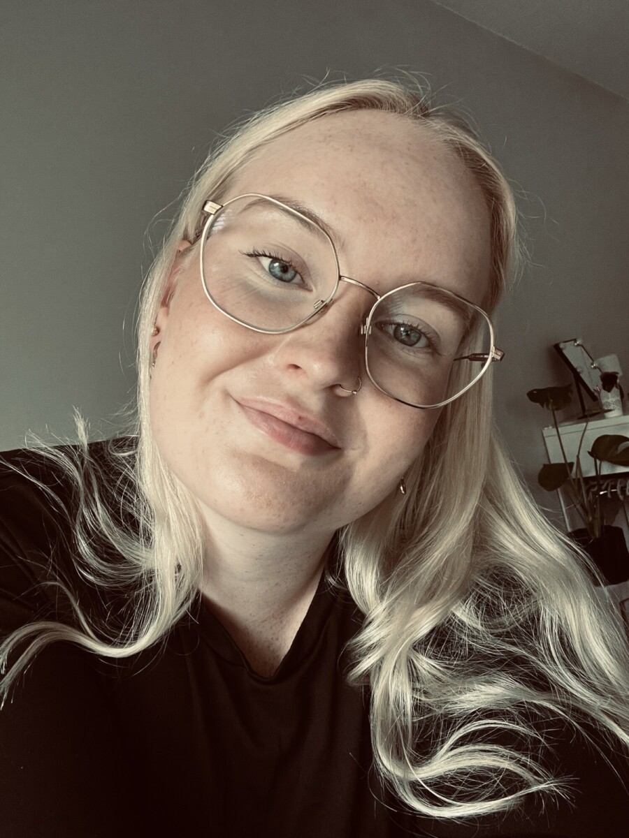 Henkilö, jolla on pitkät vaaleat hiukset ja suuret pyöreät silmälasit, hymyilee lempeästi kameralle. Hänellä on yllään musta toppi ja hän on sisätiloissa, joissa pehmeässä taustavalossa näkyvät hyllyt ja kasvi.