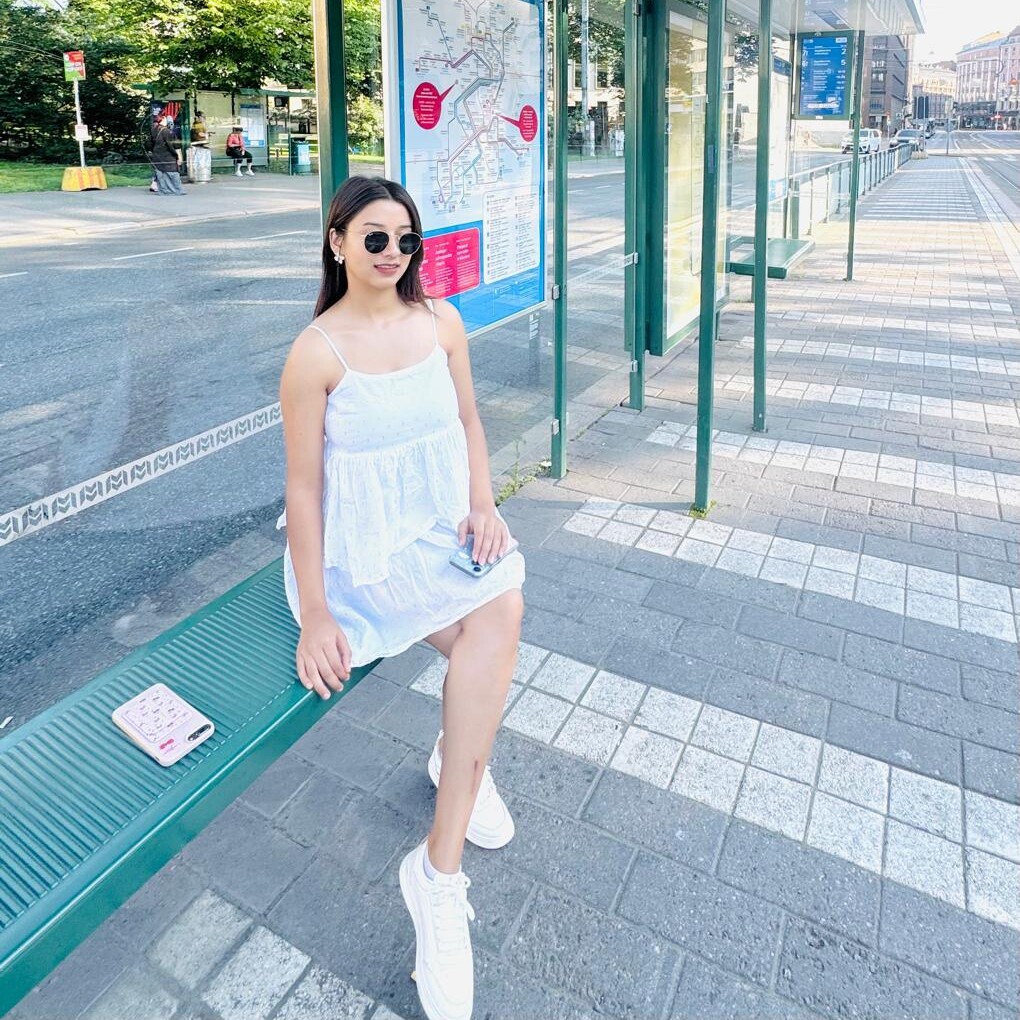 Valkoiseen mekkoon ja aurinkolaseihin pukeutunut nainen istuu vihreällä penkillä aurinkoisella bussipysäkillä, ja hänen takanaan on kaupungin kartta ja kadun toisella puolella näkyy puita.