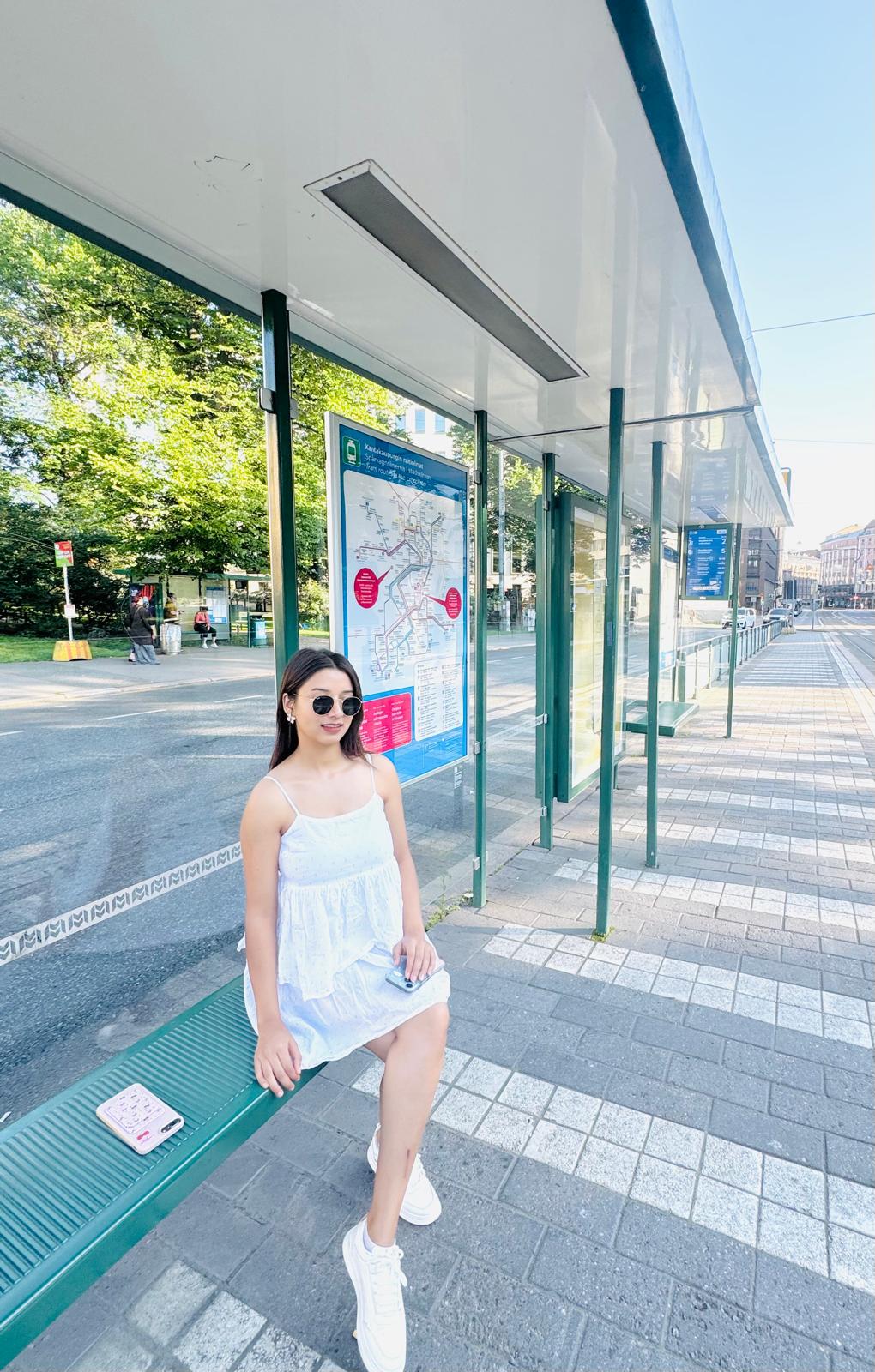 Valkoiseen mekkoon ja aurinkolaseihin pukeutunut nainen istuu vihreällä penkillä aurinkoisella bussipysäkillä, ja hänen takanaan on kaupungin kartta ja kadun toisella puolella näkyy puita.