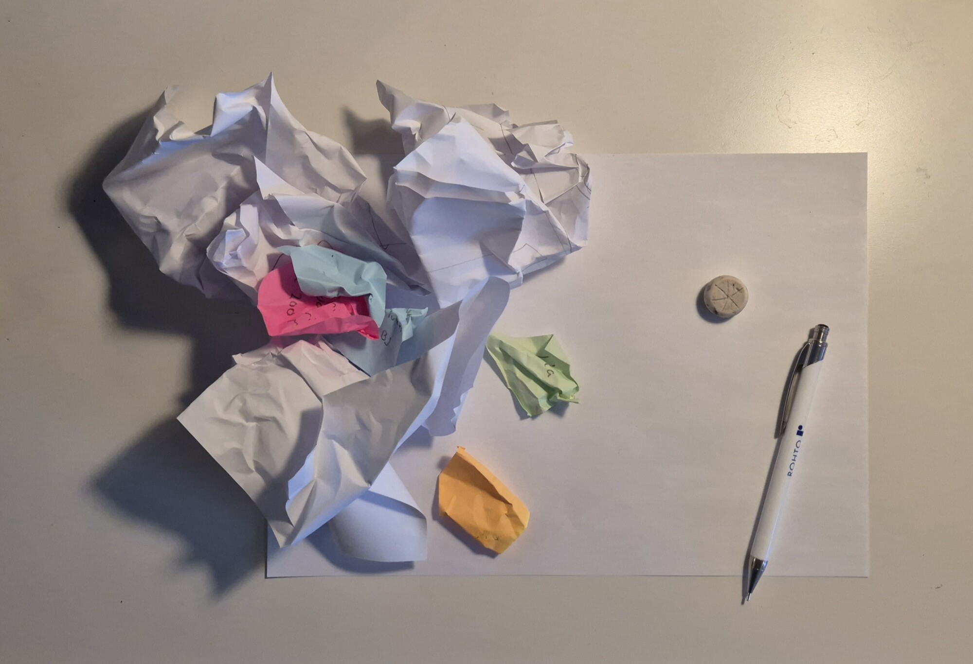 Valkoisten ja värillisten papereiden rypistynyt kasa on tyhjän arkin päällä, pienen pyyhekumin ja valkoisen lyijykynän vieressä vaalealla pöydällä.