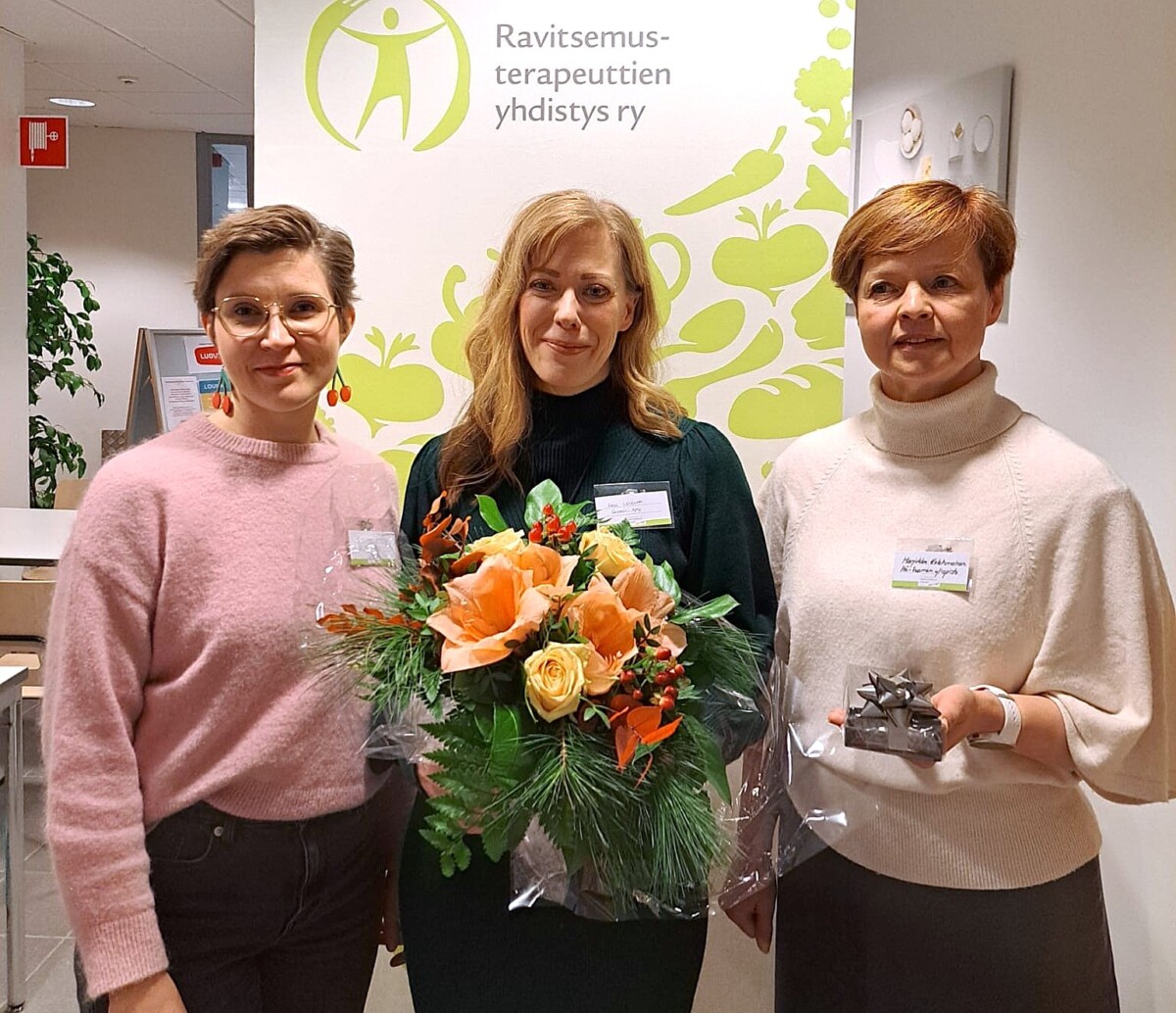 Kolme naista seisoo sisätiloissa, kahdella on kädessään lahjat - yhdellä on oranssin värinen kukkakimppu, toisella pieni kääritty laatikko. He hymyilevät Ravitsemusterapeuttien yhdistys ry:n kyltin edessä.