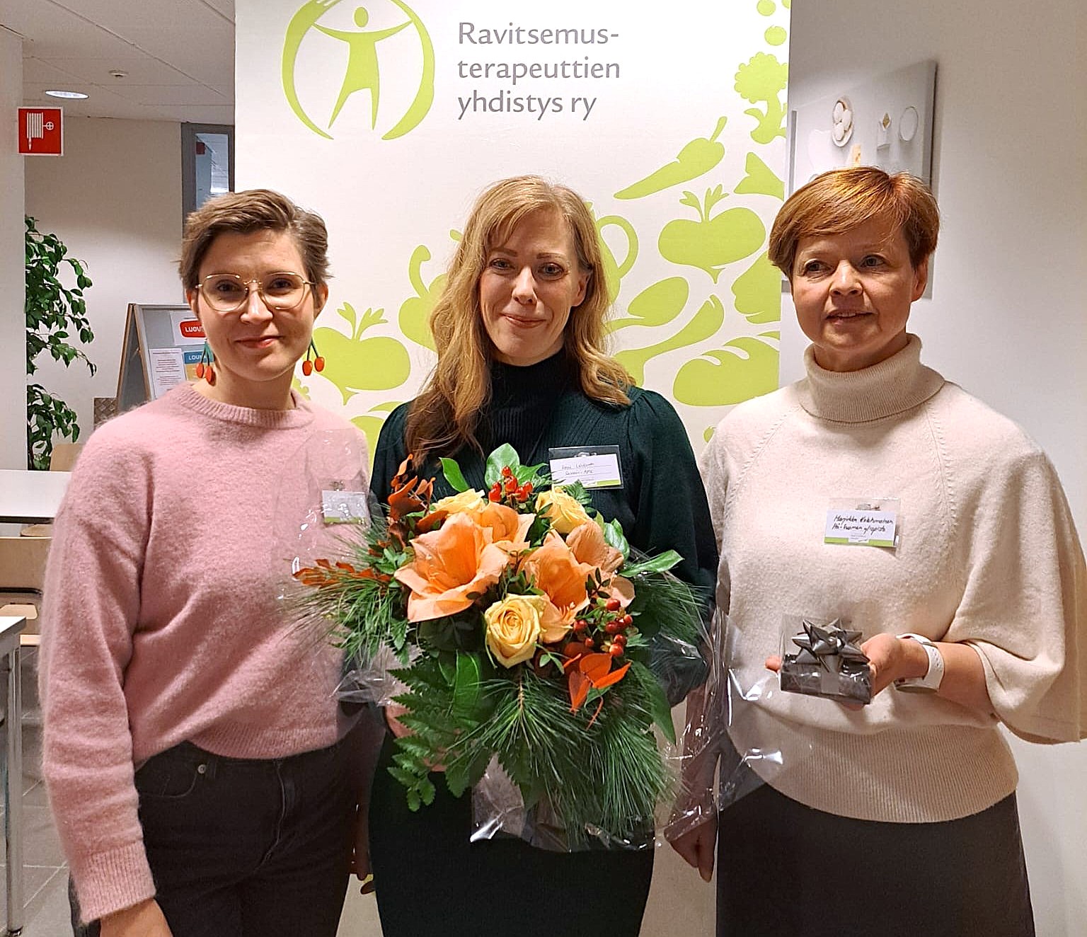 Kolme naista seisoo sisätiloissa, kahdella on kädessään lahjat - yhdellä on oranssin värinen kukkakimppu, toisella pieni kääritty laatikko. He hymyilevät Ravitsemusterapeuttien yhdistys ry:n kyltin edessä.