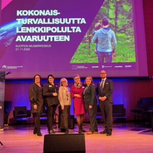 Kuusi ihmistä poseeraa ja hymyilee lavalla suuren näytön edessä, jossa on suomenkielinen teksti ja kuva perheestä luonnossa. Ympäristö näyttää olevan konferenssi tai tapahtuma Kuopion Musiikkikeskuksessa.