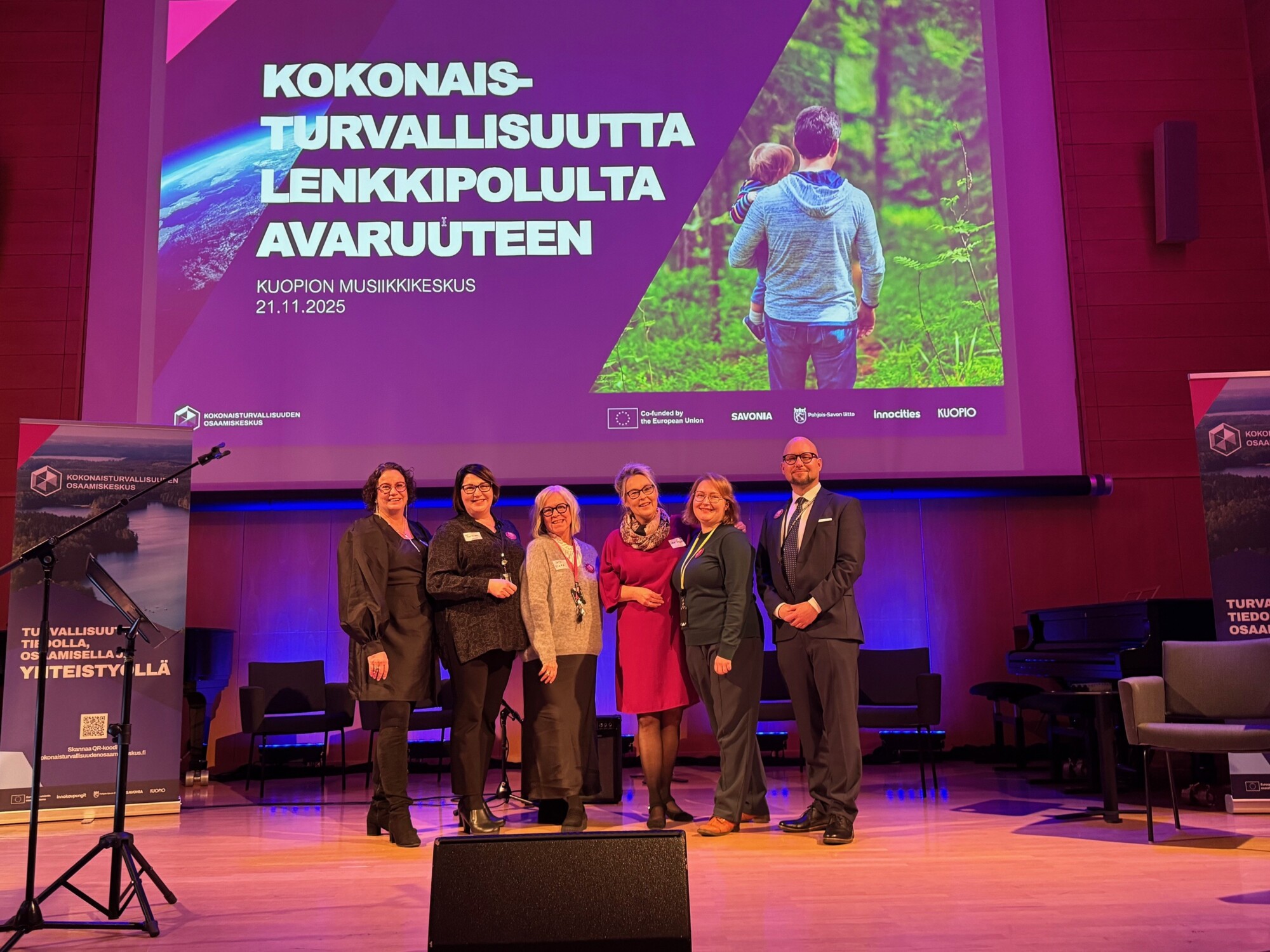 Kuusi ihmistä poseeraa ja hymyilee lavalla suuren näytön edessä, jossa on suomenkielinen teksti ja kuva perheestä luonnossa. Ympäristö näyttää olevan konferenssi tai tapahtuma Kuopion Musiikkikeskuksessa.