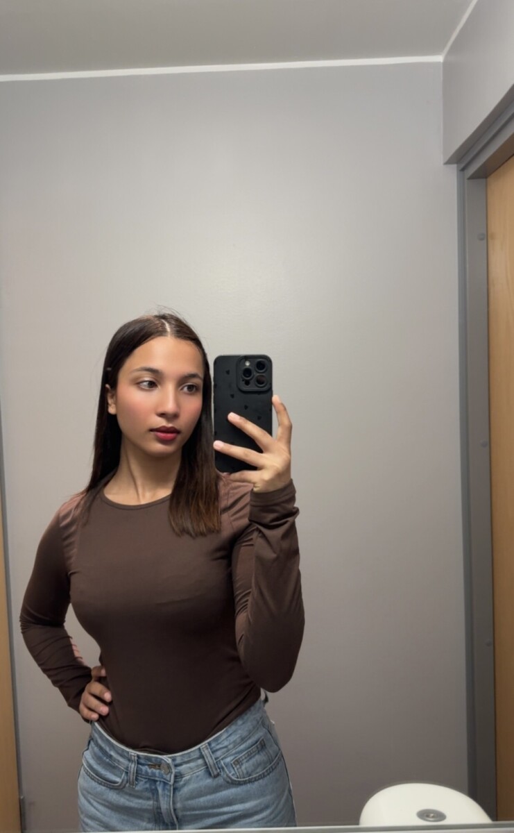 Nuori nainen, jolla on suorat ruskeat hiukset, ottaa peiliselfien kylpyhuoneessa. Hänellä on yllään pitkähihainen ruskea toppi ja siniset farkut, ja hän pitää oikeassa kädessään mustaa puhelinta. Taustalla on vaaleanharmaat seinät ja puinen ovi.