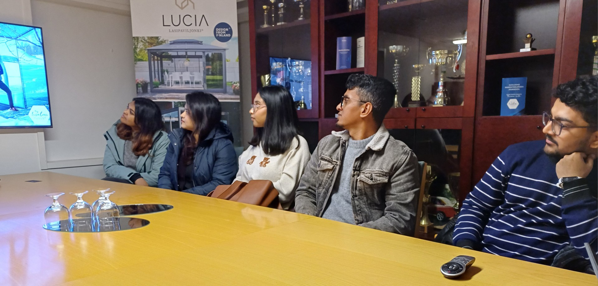 Viisi ihmistä istuu neuvottelupöydän ääressä ja katsoo tarkkaavaisesti vasemmalla olevaa näyttöä. Taustalla näkyy hyllyjä, joissa on pokaaleja ja Lucia-banneri. Etualalla pöydällä istuu kolme tyhjää lasia.