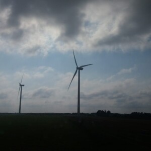 Kolme tuulivoimalaa seisoo avoimella vihreällä pellolla pilvisen taivaan alla, ja pilvien läpi pilkistää auringonvaloa. Maisema näyttää rauhalliselta ja avaralta.