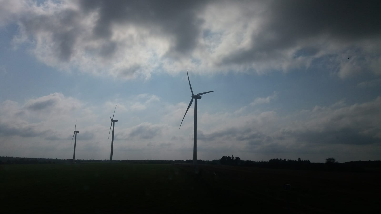 Kolme tuulivoimalaa seisoo avoimella vihreällä pellolla pilvisen taivaan alla, ja pilvien läpi pilkistää auringonvaloa. Maisema näyttää rauhalliselta ja avaralta.