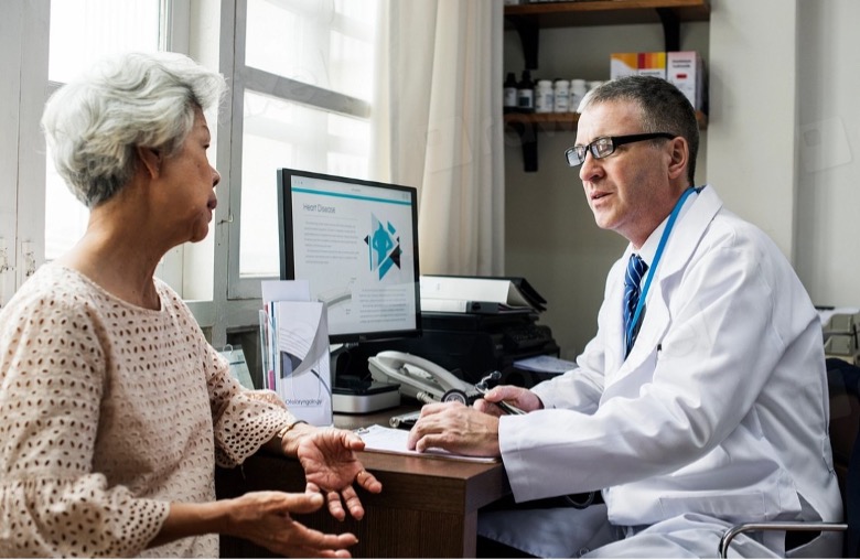 Vanhempi nainen keskustelee mieslääkärin kanssa toimistossa. Lääkäri istuu työpöydän ääressä tietokoneen ääressä, jossa näkyy sairauskortti, ja he näyttävät keskustelevan vakavasti naisen terveydestä.