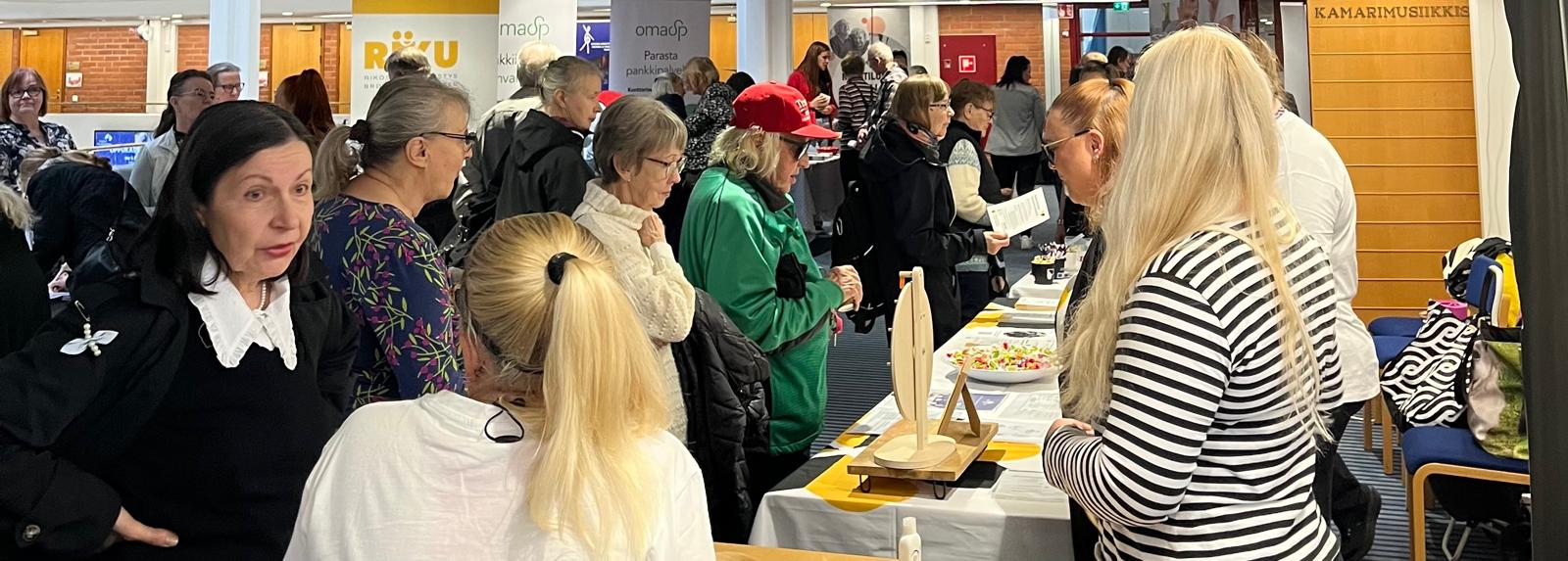 Ryhmä ihmisiä kerääntyy pöytien ympärille vilkkaassa sisätiloissa järjestettävässä tapahtumassa tai messuilla, keskustelevat ja selaavat infonäyttöjä ja -materiaaleja. Joillakin on nimilaput ja rento pukeutuminen. Taustalla näkyy kojuja ja banderolleja.