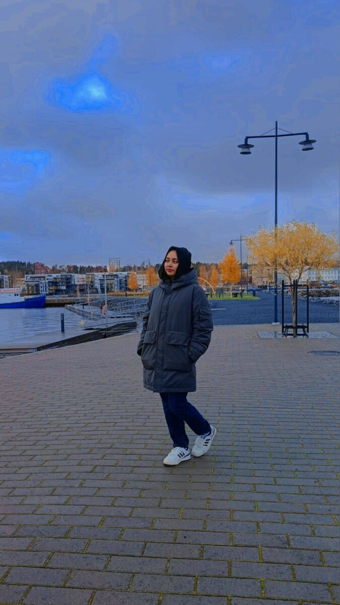 Harmaaseen takkiin, sinisiin farkkuihin ja valkoisiin lenkkareihin pukeutunut nainen seisoo päällystetyllä alueella veden äärellä, ja taustalla näkyy syksyisiä puita ja rakennuksia pilvisen sinisen taivaan alla.