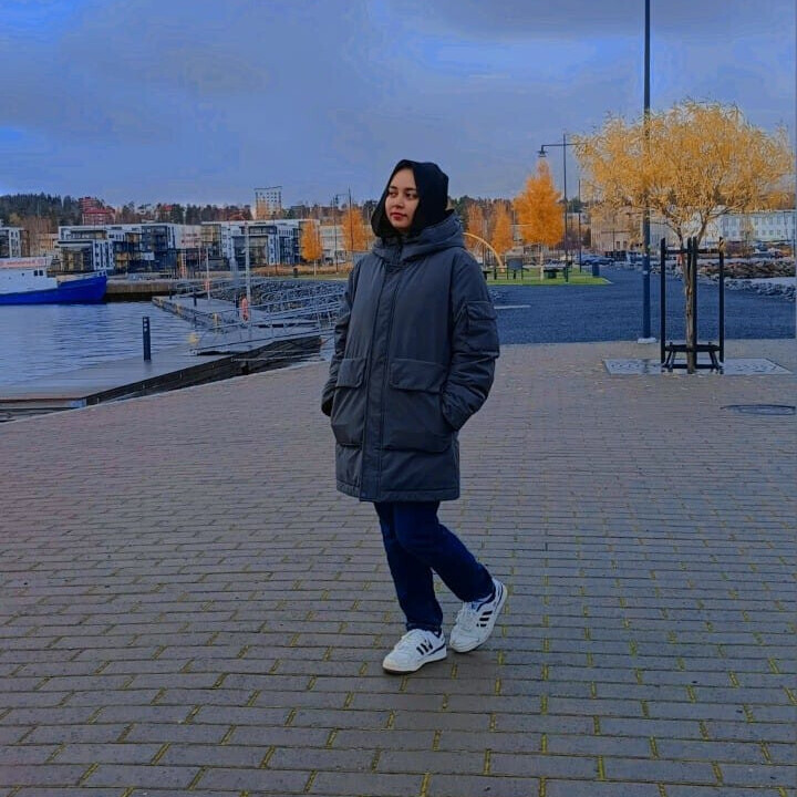 Harmaaseen takkiin, sinisiin farkkuihin ja valkoisiin lenkkareihin pukeutunut nainen seisoo päällystetyllä alueella veden äärellä, ja taustalla näkyy syksyisiä puita ja rakennuksia pilvisen sinisen taivaan alla.