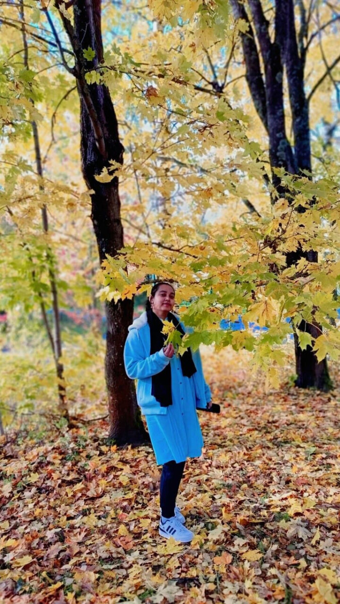 Siniseen takkiin ja mustaan huiviin pukeutunut nainen seisoo syksyn keltaisten lehtien peittämien puiden keskellä hymyillen ja oksa kädessään. Maa on pudonneiden lehtien peitossa. Kohtaus on valoisa ja värikäs.