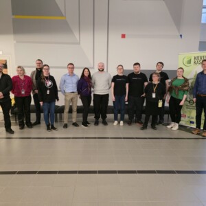 Viidentoista henkilön ryhmä poseeraa yhdessä sisätiloissa valoisassa, modernissa tilassa. Jotkut seisovat, jotkut ovat hieman kyyryssä. Oikealla on vihreä banneri, jossa on tekstiä, ja taustalla näkyy ovi ja ikkunoita.