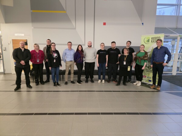 Viidentoista henkilön ryhmä poseeraa yhdessä sisätiloissa valoisassa, modernissa tilassa. Jotkut seisovat, jotkut ovat hieman kyyryssä. Oikealla on vihreä banneri, jossa on tekstiä, ja taustalla näkyy ovi ja ikkunoita.