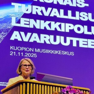 Silmälasipäinen nainen puhuu kukkien koristelemalla korokkeella. Hänen takanaan on suuri violetti näyttö, jossa on valkoinen suomenkielinen teksti ja päivämäärä 21.11.2025, joka viittaa Kuopion Musiikkikeskuksen tapahtumaan.