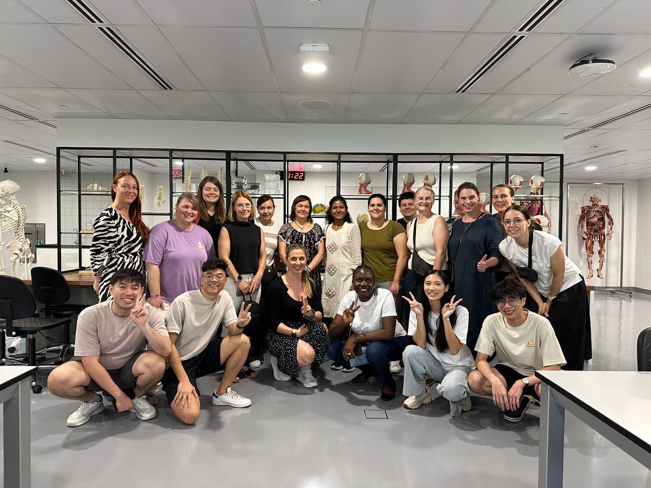 Kuusitoista ihmistä poseeraa ja hymyilee valokuvalle modernissa luokkahuoneessa, jonka taustalla on anatomisia malleja ja luurankoja. Ryhmä on monipuolinen, osa ihmisistä seisoo ja osa polvistuu tai istuu edessä.