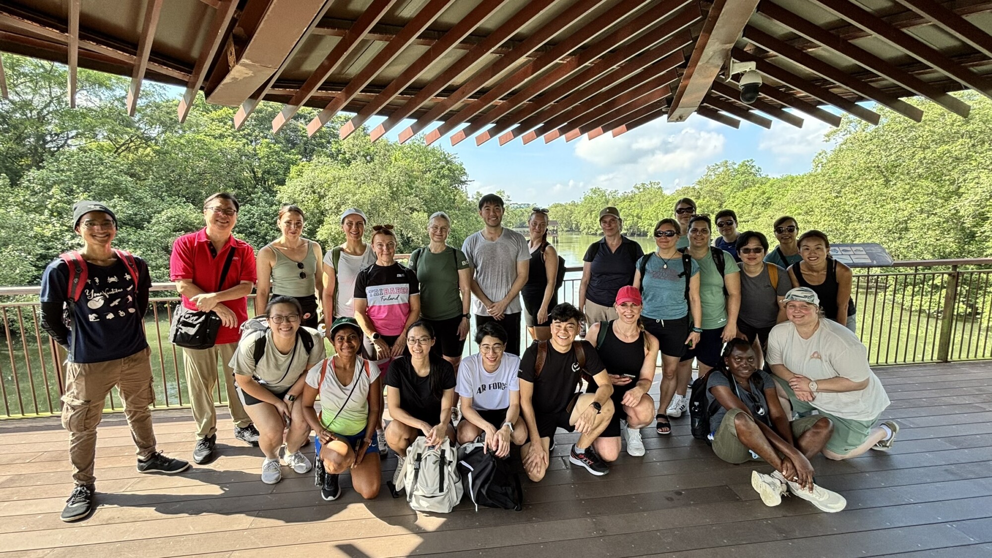 Noin 25 hengen ryhmä poseeraa ja hymyilee yhdessä puisella kannella paviljongin alla, taustalla puita ja vehreyttä aurinkoisena päivänä.