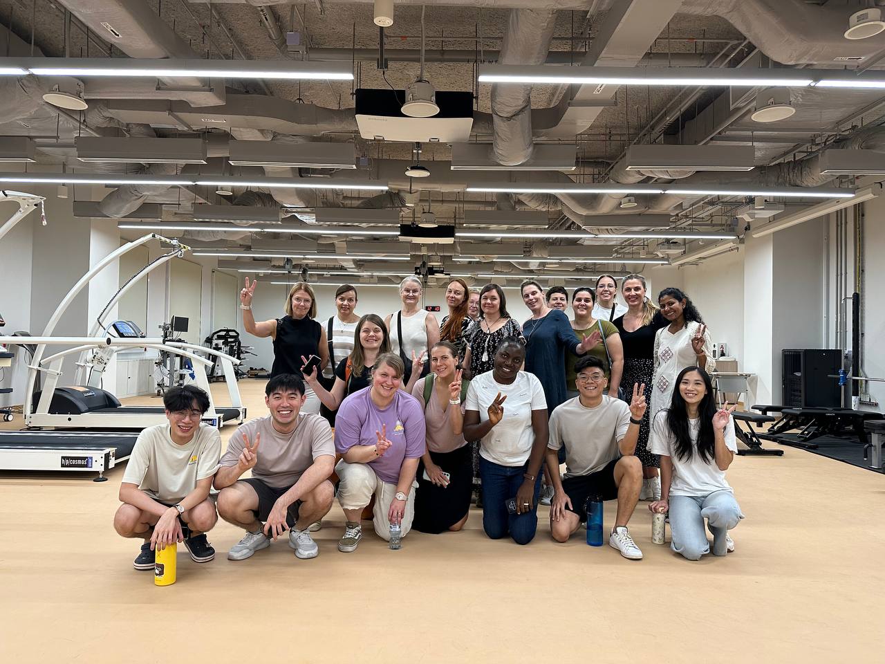 Erilaiset ihmiset poseeraavat yhdessä kuntosalilla hymyillen ja rauhanmerkkejä tehden. Taustalla näkyy liikuntavälineitä ja juoksumattoja. Ympäristö on hyvin valaistu ja siinä on teollinen katto.