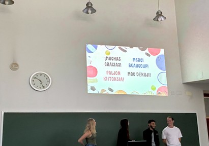 Neljä ihmistä seisoo luokkahuoneessa liitutaulun ja valkokankaan edessä. Näytöllä näkyy Kiitos useilla kielillä, ja kello näyttää kello 10:13. Näytöllä näkyy urheiluvälinekuvakkeita.